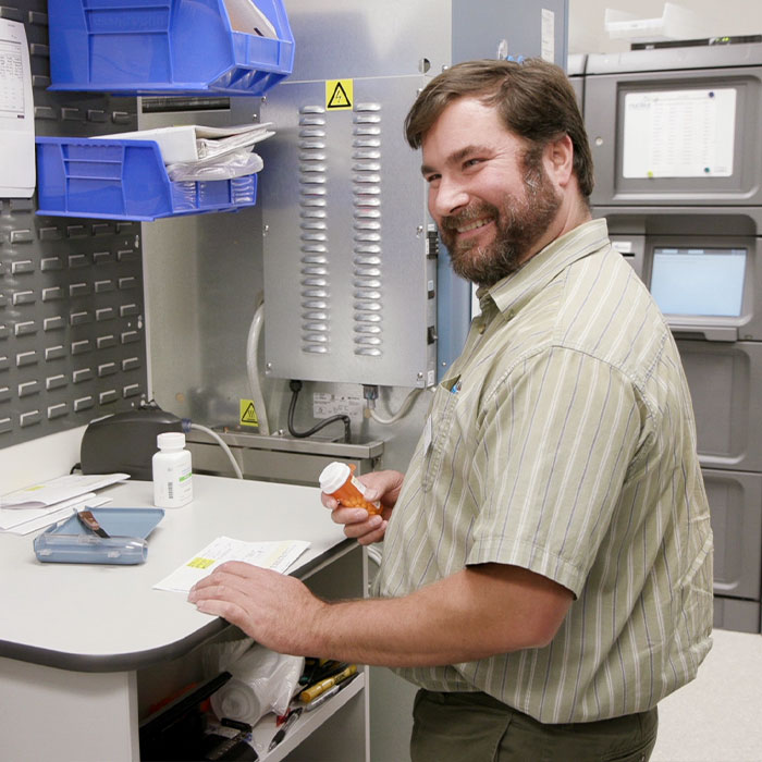 An oncology pharmacy technician holds a pill bottle and smiles over his shoulder towards the camera
