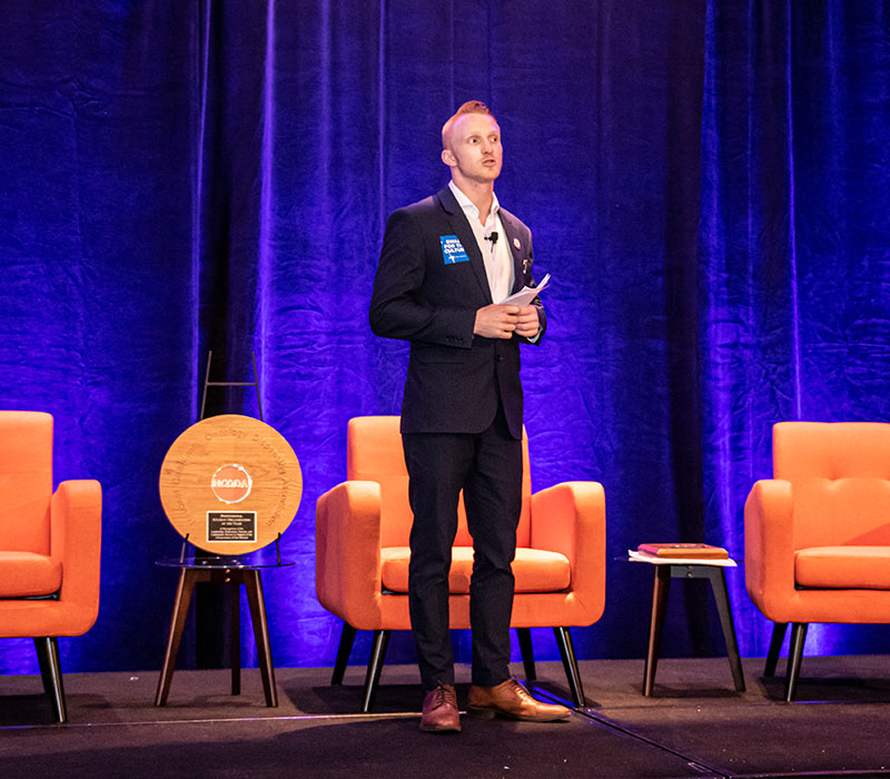 A previous NCODA fellow stands on a stage in front of orange chairs, giving a presentation